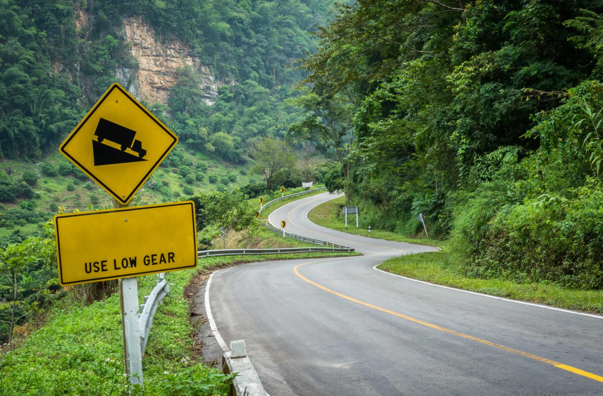 Estrada sinuosa em uma curva acentuada, com uma placa de trânsito à margem exibindo o desenho de um caminhão e a mensagem “Reduza a velocidade”, alertando motoristas sobre o risco de acidentes e a necessidade de cautela, especialmente para veículos pesados. Estrada sinuosa em uma curva acentuada, com uma placa de trânsito à margem exibindo o desenho de um caminhão e a mensagem “Reduza a velocidade”, alertando motoristas sobre o risco de acidentes e a necessidade de cautela, especialmente para veículos pesados.