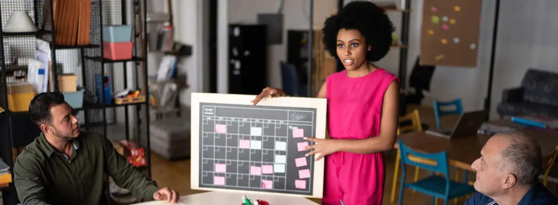 Mulher segura quadro com calendário e post-its, em uma reunião com equipe na gestão de projetos. Mulher segura quadro com calendário e post-its, em uma reunião com equipe na gestão de projetos.